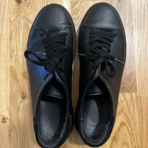 Svarta sneakers från Axel Arigato strl 39 - Snygga svarta sneakers från Axel Arigato med klassisk design och svarta snören. Skorna har rund tå, platt sula och är tillverkade i skinn. Perfekta för en stilren och minimalistisk look. I använt men mycket fint skick! 