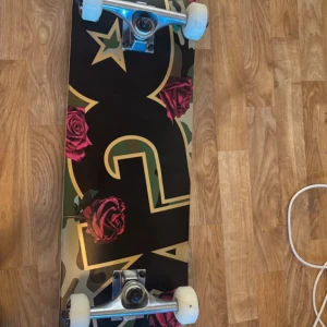 Skateboard med DC-motiv och rosor! - Säljer en skateboard med svart griptape på ovansidan och en snygg undersida med DC-logga, kamouflagemönster och röda rosor. Brädan har vita hjul och silverfärgade trucks. Perfekt för dig som gillar streetstyle och unika designer.