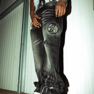 Svarta wide jeans med tryck och slitningar - Säljer ett par svarta jeans med bred passform och unikt tryck på benen. Jeansen har slitna detaljer och ett stort tryckt märke på låret. 