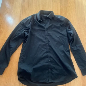 Svart skjorta från Dressmann - Klassisk svart skjorta från Dressmann i bomullsblandning. Skjortan har lång ärm, knappar framtill och en stilren krage. Perfekt för dig som gillar en enkel och stilren look.