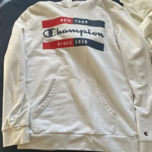 Vit hoodie från Champion med tryck - Säljer en vit hoodie från Champion med tryck i rött, blått och svart framtill. Tröjan har känguruficka och långa ärmar. Klassisk och sportig stil med broderad logga på ärmen.