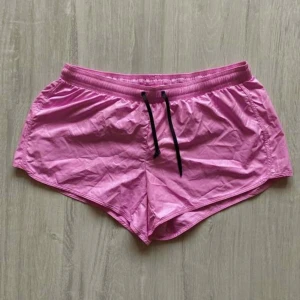 Rosa sportshorts från H&M - Säljer ett par rosa sportshorts från H&M med svart dragsko i midjan. Shortsen har en klassisk sportig design och är tillverkade i ett lätt syntetmaterial som torkar snabbt. Perfekta för träning eller löpning.