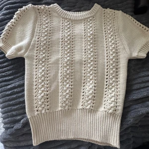 Beige stickad topp från Zara - Säljer en beige stickad topp från Zara med korta ärmar och vackert hålmönster. Tröjan har ribbad kant vid hals, ärmslut och nederkant. Perfekt för lager-på-lager eller sommarens svalare dagar.