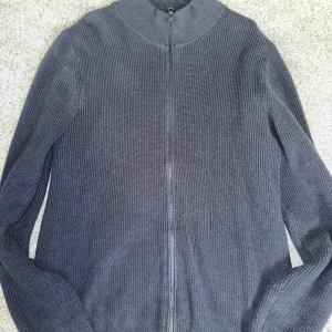 Mörkblå stickad kofta med dragkedja från John Henric - Säljer en mörkgrå stickad kofta med hel dragkedja framtill och hög krage. Tröjan har lång ärm och är ribbstickad för en stilren look. Perfekt för lager-på-lager under kyliga dagar. Trendigt tröja från John Henric 