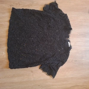 Svart spetsblus med kort ärm - Säljer en svart blus i spets med korta ärmar och fina spetsdetaljer. Blusen har rund halsringning och ett elegant mönster i tyget. Perfekt för dig som vill ha en stilren och trendig topp.