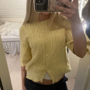 Gul stickad kofta  - Söt ljusgul stickad kofta med korta ärmar och knappar framtill. Koftan har ett fint kabelstickat mönster och ribbade kanter vid ärmslut, hals och nederkant. Perfekt att slänga över en topp för en fräsch look.