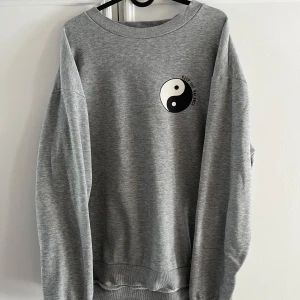 Grå sweatshirt med yin yang-tryck från 157 - Grå sweatshirt från 157 i storlek M med ett svartvitt yin yang-tryck och texten 'Keep the balance' på bröstet. Tröjan har rund halsringning och långa ärmar. Mjuk insida och klassisk passform. Har lite lösa trådar i armslutet men inget som syns på utsidan