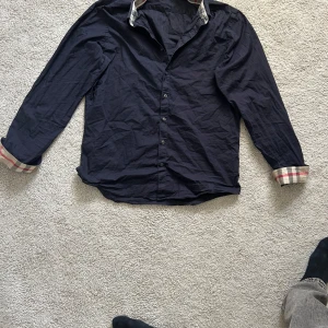 Mörkblå skjorta med rutiga detaljer från Burberry - Säljer en mörkblå skjorta från Burberry med klassiska rutiga detaljer på insidan av kragen och ärmsluten. Skjortan har knappar framtill och lång ärm. Perfekt för dig som gillar stilrena plagg med en twist.