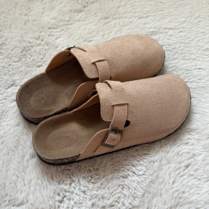 Beige slip-in sandaler med spänne - Säljer ett par beige slip-in sandaler med justerbart metallspänne och mockaliknande ovandel. Skorna har en platt sula med korkdetalj på sidan och svart undersida. Perfekta för en avslappnad stil.
