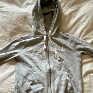 Grå hoodie från Bondelid - Mjuk och skön grå hoodie från Bondelid med vit broderad text och siffrorna 1956 på bröstet. Tröjan har dragkedja, huva med snören och två stora fickor framtill. Perfekt för en avslappnad stil.