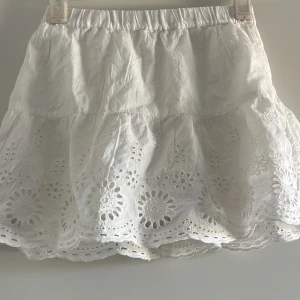 Vit broderad kortkjol med volang - Superfin vit kjol med broderade blommönster och volangkant. Kjolen har resår i midjan och är i en luftig bomullskvalitet. Perfekt för varma dagar och ger en söt och somrig look.