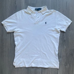 Vit pikétröja från Polo Ralph Lauren - Klassisk vit pikétröja från Polo Ralph Lauren med broderad marinblå logga på bröstet. Tröjan har korta ärmar, krage och knappar framtill. Tillverkad i 100% bomull för en skön och luftig känsla. Släppt lite i sömnen längst ner men inget som märks när man bär den