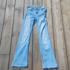 Ljusblå bootcut jeans - Säljer ett par ljusblå jeans med bootcut passform och hög midja. Klassisk femficksmodell med dragkedja och knapp framtill. Jeansen har en liten lila fläck och ett hål på ena benet. Perfekta för dig som gillar vintage vibe.