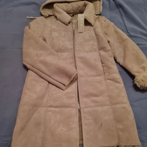 Beige lång kappa från Ytko Jeans - Säljer en beige lång kappa från Ytko Jeans med mjuk, pälsliknande insida och dragkedja framtill. Jackan har klassisk krage och långa ärmar, perfekt för kyligare dagar. Snygg och stilren design som passar till många outfits.