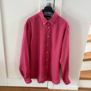 Rosa linneskjorta från Tommy Hilfiger - Snygg rosa skjorta från Tommy Hilfiger i 100% linne. Klassisk button-down krage, vita knappar och diskret logga på bröstet. Perfekt för dig som vill ha en stilren och luftig skjorta med lång ärm.