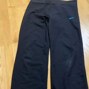 Svarta mjukisbyxor från Nike - Svarta mjukisbyxor från Nike med blå broderad logga framtill. Byxorna har en normal passform och är tillverkade i mjukt bomullsmaterial. Perfekta för en avslappnad stil.