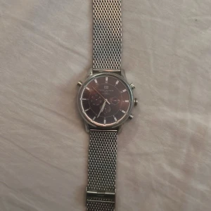 Tommy Hilfigher Silverfärgad kronografklocka   - Stilren herrklocka från Tommy Hilfigher med svart urtavla och silverfärgat mesharmband i metall. Klockan har tre små urtavlor och tydliga markeringar. Perfekt accessoar för dig som gillar klassisk design.