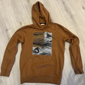 Brun hoodie med tryck från Kappahl - Säljer en brun hoodie från Kappahl med coolt skateboard-tryck på bröstet. Tröjan har huva och långa ärmar, perfekt för en avslappnad stil. Skön bomullskänsla och passar dig som gillar streetwear.