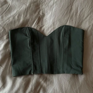 Mörkgrön korsett topp - Säljer en mörkgrön korsettopp med markerade sömmar och hjärtformad urringning. Toppen är axelbandslös och har smockad rygg samt dragkedja i sidan. En liten defekt, de bild 3