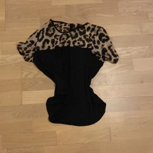 Svart topp med leopardmönstrade axlar - Säljer en svart topp med korta ärmar och leopardmönstrad överdel. Toppen har rund halsringning och är figurnära i modellen. Perfekt för dig som vill sticka ut lite extra med ett djurmönster.
