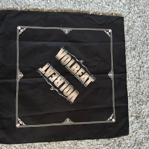 Svart halsduk med Volbeat-tryck 55 x 55 cm - Cool svart halsduk med Volbeat-logga i mitten och dekorativa vita ramar runtom. Perfekt för fans av bandet som vill visa sitt stöd på konserter eller i vardagen. Mått 55 x 55 cm. Helt ny. Bomull