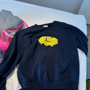 Svart sweatshirt med gul vintage-tryck - Svart sweatshirt med ett stort gult vintage-tryck på bröstet. Klassisk rund hals och långa ärmar. Perfekt för en avslappnad stil.
