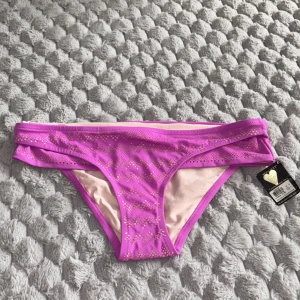 Lila bikiniunderdel från Victoria's Secret - Säljer en lila bikiniunderdel från Victoria's Secret med små glittriga detaljer och snyggt band upptill. Materialet är syntet och insidan är ljusrosa. Perfekt för stranden eller poolen!