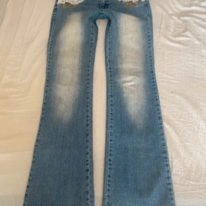 Blå bootcut jeans med spetsdetaljer - Säljer ett par ljusblå bootcut jeans med vit spetsdekor framtill och på bakfickorna. Midjemätt:38-39cm Grenmätt:20cm Innerbenslängd:83cm Benöppning:21cm Inga defekter!