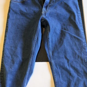 Blå jeansbyxor från GANT med regular fit - Säljer ett par klassiska blå jeans från GANT med regular fit och raka ben. Byxorna har midwaist, fem fickor och snygga detaljer som GANT-lapp i läder bak. Perfekta för en stilren och avslappnad look.