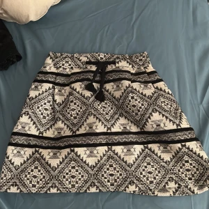 Svartvit kjol med grafiskt mönster och knytband - Säljer en svartvit kjol med grafiskt mönster och knytband i midjan. Kjolen har en A-linjeform och är i kort modell. Perfekt för dig som gillar unika detaljer och mönster.