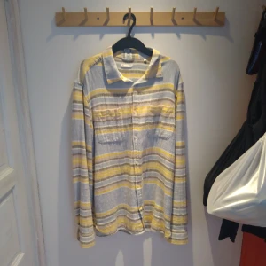 Randig skjorta från Knowledge Cotton Apparel - Säljer en långärmad skjorta från Knowledge Cotton Apparel i storlek XL. Skjortan har ett randigt mönster i ljusblå, vitt och gult, klassisk krage och knäppning framtill. Perfekt för dig som gillar en avslappnad men stilren look.