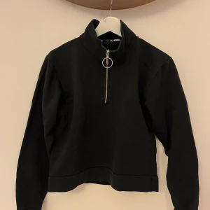 Svart halvzip tröja med ringdetalj - Svart långärmad tröja med hög krage och halv dragkedja med snygg ringdetalj. Tröjan är i bomullsblandning och har en enkel, stilren design som passar till många outfits. Passar dom som i vanliga fall är XS/S