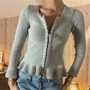 Ljusgrå stickad kofta med volangkant - Supersöt ljusgrå stickad kofta från Odd Molly med volangkant nertill och vid ärmslut. Koftan har lång ärm, dekorativa knappar framtill och en fin strukturstickning. Perfekt att slänga över en topp för en mysig look. Koftan är i storlek S och i ett mycket bra skick. 