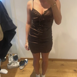 Brun tight kortklänning med smala axelband - Säljer en brun tight kortklänning med smala axelband och rynkade detaljer framtill. Klänningen har en v-ringad byst och figurnära passform som framhäver siluetten. Perfekt för dig som vill ha en stilren och trendig look.
