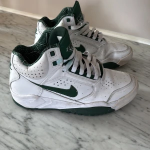 Nike Air Flight vita och gröna sneakers - Knappt använda! Nike Air Flight sneakers i vitt och grönt med högt skaft och klassisk snörning. Skorna har gröna detaljer, perforerade sidor och Nike-logga på hälen. Perfekta för dig som gillar retrostil och basketinspirerade sneakers.