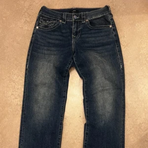 Mörkblå jeans från True Religion - Säljer ett par mörkblå jeans från True Religion med kontrastsömmar och fickdetaljer med knappar bak. Modellen är relaxed fit och har normal passform. 