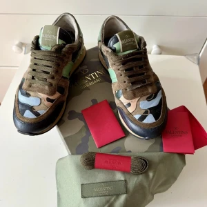 Valentino Garavani sneakers med camouflage - Säljer ett par Valentino Garavani sneakers med klassiskt camouflage-mönster i grönt, beige, svart och blått. Skorna har bruna mockadetaljer, snörning och platt sula. Kommer med originalkartong, extra skosnören och dustbag.