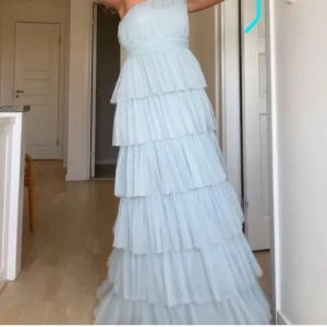 Ljusblå långklänning med volanger - Säljer en drömmig ljusblå långklänning med flera lager volanger och oneshoulder-modell. Klänningen har en romantisk känsla och är sydd i ett tunt, skirt material som ger ett svävande intryck.