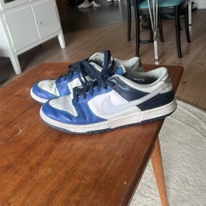 Nike dunk low sneakers - Säljer ett par Nike Dunk Low sneakers i blått och vitt med svarta skosnören. Skorna har en klassisk låg siluett och rund tå. Perfekta för dig som gillar streetwear och vill ha en ikonisk sneaker i garderoben.storlek 45💗( rengör innan jag skickar)