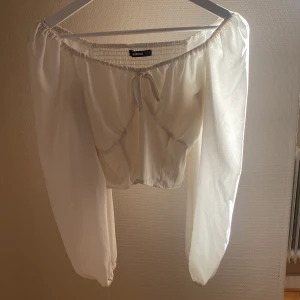 Vit offshoulder blus med knyt - Säljer en söt vit offshoulder blus från Bik Bok med långa, luftiga ärmar och knyt framtill. Blusen har smockad rygg och är perfekt för en somrig look. Tunn och lätt i materialet.