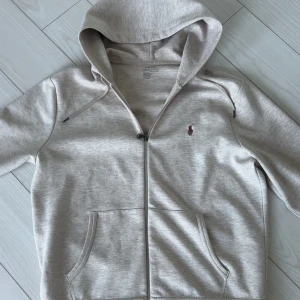 Beige hoodie från Polo Ralph Lauren - Snygg beige hoodie från Polo Ralph Lauren med dragkedja och broderad logga på bröstet. Tröjan har huva med dragsko och två fickor framtill. Perfekt för en avslappnad stil.