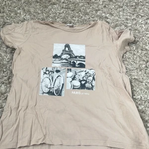 Beige t-shirt med Paris-motiv från Amisu - Säljer en beige t-shirt från Amisu med tryck av Eiffeltornet, cykel och macarons. T-shirten har korta ärmar och en avslappnad passform. Perfekt för dig som gillar Paris-vibbar och snygga tryck!