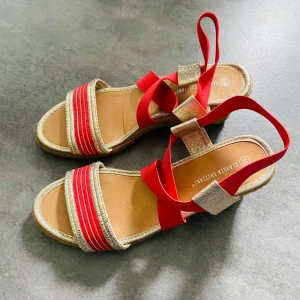 Mörk rosa/guldiga  sandaler med kilklack från Claudia Ghizzani - Snygga sandaler med mörk rosa och guldig rem, elastiska band och flätad kilklack. Öppen tå och bekväm passform, perfekta för varma dagar. Sula i naturfärgat material och detaljer i syntet. Skorna är i nyskick, möjligtvis använda en gång. 