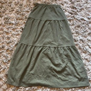 Grön kjol - Säljer en grön kjol med volangdetaljer. Har en längre modell med flera lager som ger en avslappnad och trendig look. Perfekt att styla med en enkel topp eller stickad tröja.