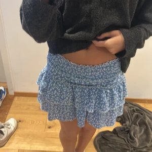 Blå volangkjol med blommigt mönster - Söt blå kjol med vitt blommigt mönster och volanger. Kjolen har resår i midjan och är kort i modellen. Perfekt för en somrig look.