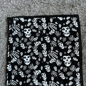 Svart halsduk med döskallemönster 50 x 50 cm - Cool svart halsduk med vita döskallar och skelett-händer som mönster. Perfekt accessoar för dig som gillar alternativ stil eller vill sticka ut lite extra. Fyrkantig form och tunnare material. Bomull. Helt ny. Mått 50 x 50 cm,