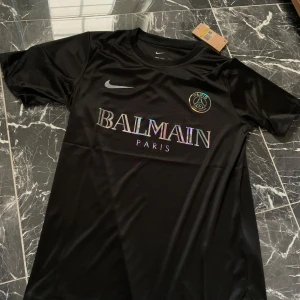 Svart PSG x Balmain fotbollströja från Nike - Säljer en svart fotbollströja från Nike i samarbete med PSG och Balmain. Tröjan har korta ärmar, rund hals och glänsande Balmain Paris-tryck på bröstet samt PSG-logga. Materialet är slätt och sportigt, perfekt för fotboll eller streetwear.