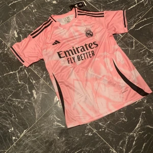 Rosa Real Madrid fotbollströja från Adidas - Säljer en rosa Real Madrid fotbollströja från Adidas med svarta detaljer och klubbmärke på bröstet. Tröjan har korta ärmar, svart Adidas-logga och tre ränder på axlarna. Perfekt för dig som vill sticka ut på planen.