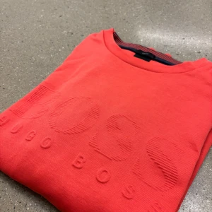 Korallröd sweatshirt från Hugo Boss - Säljer en snygg korallröd sweatshirt från Hugo Boss med diskret broderad logga framtill. Tröjan har rund halsringning och smal passform. Perfekt för dig som gillar stilrena och bekväma plagg.