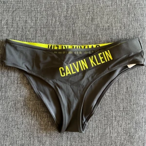 Grå bikiniunderdel från Calvin Klein - Snygg grå OANVÄND bikiniunderdel från Calvin Klein med bred resår och gul logga. 